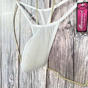 May include: White lingerie with a sheer mesh front and thin straps. The garment is displayed on a gold-colored hanger against a wooden background. A tag with the text "jovanadesign" is visible.
