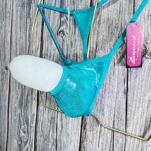 May include: A turquoise thong-style bikini bottom with a metallic sheen, displayed on a white mannequin. The garment is suspended on a gold-colored wire hanger. A tag with the brand name "Jovanadesign" is attached.