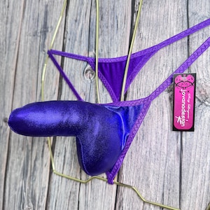 May include: A purple metallic thong-style garment with a pouch. The lingerie is displayed on a gold-colored hanger against a wooden background. A tag with the text "Jovandesign" is attached.