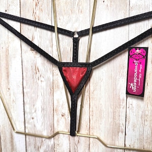 Pode incluir: Uma tanga preta com um painel triangular central em vermelho metálico, alças elásticas pretas finas e um pequeno anel de metal. Uma etiqueta retangular presa na lateral apresenta o texto jovanadesign em branco sobre um fundo rosa e preto.
