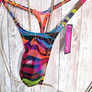 May include: A thong featuring a vibrant, multi-colored animal print pattern in neon orange, pink, blue, and green with a black dot texture. The garment includes thin straps, gold-tone metal rings, and a tag labeled jovanadesign.