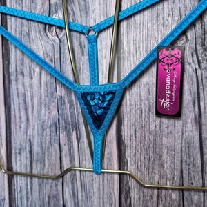 May include: A bright blue thong with thin elastic straps, a central metal ring, and a triangular front panel featuring a dark blue and black snakeskin-like pattern. A rectangular black tag with pink text reading jovanadesign is attached.