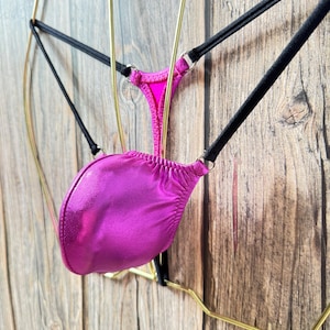 May include: A pink satin thong with black straps hanging on a gold hanger. The thong is made of a shiny, silky fabric.