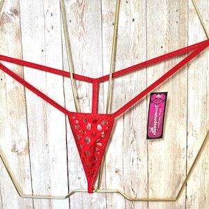 May include: A red thong featuring a geometric cutout pattern on the front panel and thin side straps. The garment includes a black rectangular tag with the text "jovanadesign" in white lettering, displayed on a gold-toned wire hanger.