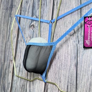 May include: A blue and black mesh thong with a decorative design, suspended on a gold-coloured hanger. The thong features a mesh pouch and blue straps. The tag on the right side reads "jovanadesign".