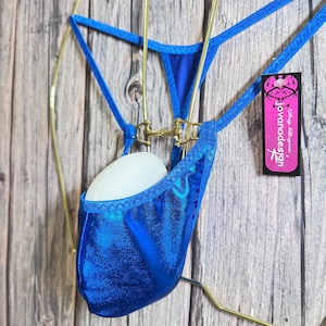 May include: A blue metallic thong swimsuit with gold-colored hardware. The swimsuit is hanging on a gold-colored hanger. A tag with the text "Jovanadesign" is attached to the swimsuit.