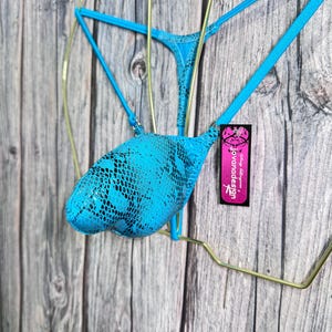 May include: A turquoise bikini set with a snakeskin pattern. The bikini is displayed on a gold-colored hanger against a wooden background. A tag with the text "jovandadesign" is attached.