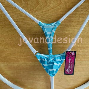 Puede incluir: Braguita de bikini tanga a rayas blancas y azul claro con tirantes blancos. La prenda se muestra sobre una percha blanca sobre un fondo de madera. Se adjunta una etiqueta con el texto "jovanadesign".