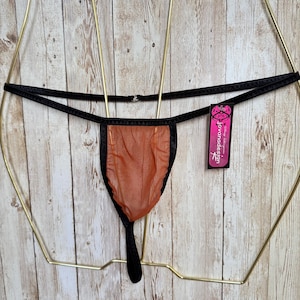 May include: A thong featuring a sheer, burnt orange mesh front panel with black trim and thin black side straps. A rectangular black tag with pink text reading "jovanadesign" is attached to the side. The garment is displayed on a gold wire frame.