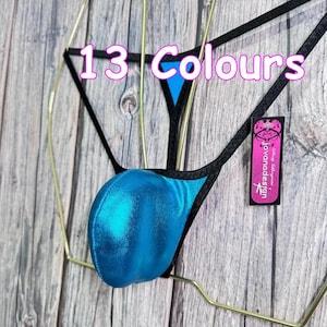 Puede incluir: Un tanga de traje de baño turquesa con ribete negro se muestra en una percha de alambre dorado sobre un fondo de madera. El texto "13 Colores" es visible. También hay una pequeña etiqueta con el texto "jovanadesign".