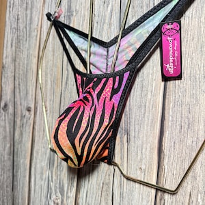 May include: A black and neon pink zebra print thong swimsuit bottom. The swimsuit has a black trim and is hanging on a gold-colored hanger. A tag with the text "jovanadesign" is attached to the hanger.