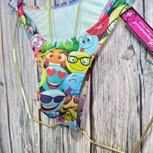 Könnte beinhalten: Bunter Bikini-Slip mit einem Emoji-Muster. Das Design umfasst verschiedene Emoji-Gesichter mit unterschiedlichen Ausdrücken, wie Sonnenbrillen, Herzen und Lächeln. Das Kleidungsstück wird auf einem goldfarbenen Kleiderbügel vor einem Holzgrund präsentiert.