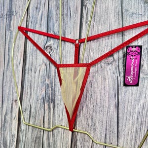 May include: Red and beige lingerie with a delicate design. The lingerie features a red trim and a beige mesh panel. The garment is displayed on a gold-colored hanger against a wooden background. A tag with the text "jovanadesign" is visible.