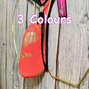 以下が含まれることがあります： ネオンコーラルのTバック。ゴールドのアクセントと黒のトリムが施され、ゴールドのハンガーに展示されています。「3 Colours」の文字が画像に重ねられています。「Jovanadesign」というブランド名のタグが見えます。