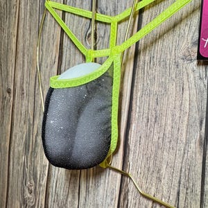 Könnte beinhalten: Ein neon-grüner Mesh-Jockstrap mit einem schwarzen Mesh-Beutel. Der Jockstrap hängt an einem goldenen Kleiderbügel vor einem Holz-Hintergrund.