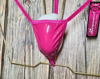 May include: A bright pink thong-style lingerie piece with a shiny, patent leather-like finish. The garment is suspended on a gold-colored metal hanger, showcasing its delicate straps and design. A tag with the text "jovanadesign" is visible.