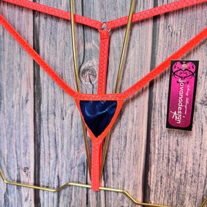 May include: A neon orange thong featuring thin elastic straps and a central dark blue metallic triangular panel. A black rectangular tag is attached to the side, displaying the white text "jovanadesign" and a stylized graphic logo design.