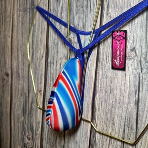 May include: Blue thong underwear featuring a red, white, and blue pattern. The garment hangs on a gold-coloured wire hanger. A tag with the text "jovanadesign" is attached.