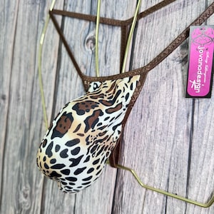 Könnte beinhalten: Ein String-Badeanzug mit Leopardenmuster und brauner Einfassung. Der Badeanzug wird auf einem goldfarbenen Kleiderbügel vor einem Holzgrund gezeigt. Ein rosa Etikett mit dem Text "Jovanadesign" ist angebracht.