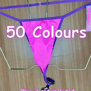 May include: A pink thong with purple trim and a purple string attached to a black plug. The thong is hanging on a metal hanger. The text "50 Colours" is written in white on a pink background. The text "Plug is not included" is written in white on a pink background.