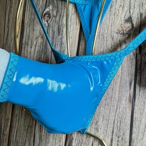 May include: Bright blue thong-style underwear with a shiny, patent leather-like finish. The edges are trimmed with a textured, patterned fabric. The garment is displayed on a gold-colored hanger against a wooden background.