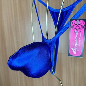 May include: A vibrant royal blue thong featuring a contoured front pouch and thin elastic straps. A rectangular black tag with pink accents is attached, displaying the text 'Sexy Lingerie' and 'jovanadesign' in white and black lettering.