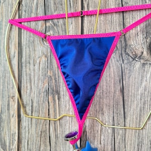 May include: A blue satin thong with pink trim hanging on a gold hanger. The thong has a small, silver-colored plug attached to the bottom.