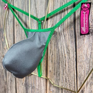 Könnte beinhalten: Grüne und graue Dessous mit einem Joviana Design-Etikett. Die Dessous hängen an einem goldenen Kleiderbügel vor einem Holz Hintergrund.