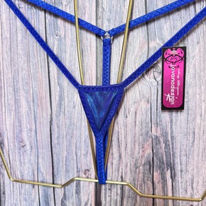 May include: A royal blue thong featuring thin elastic straps, a central triangular panel with a shimmering iridescent finish, and a small metal ring connector. A black rectangular tag is attached, displaying the text jovanadesign.