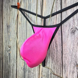 Könnte beinhalten: Rosa und schwarze Dessous mit einem dünnen Riemen-Design. Die Dessous hängen an einem goldenen Kleiderbügel.