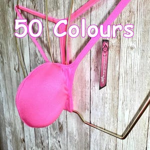 Könnte beinhalten: Ein leuchtend neonpinker Tanga mit dünnen Trägern und einer abgerundeten Fronttasche, präsentiert auf einem goldfarbenen Drahtbügel. Der Text 50 Colours ist in Weiß mit rosa Umrandung überlagert und ein schwarzes Etikett jovanadesign ist befestigt.