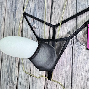 K&ouml;nnte beinhalten: Schwarzes Dessous-Set mit einem transparenten String-Tanga und einem BH-&auml;hnlichen Design. Das Set wird auf einem goldfarbenen Kleiderb&uuml;gel vor einem Holzgrund pr&auml;sentiert. Der Stoff wirkt zart und das Design ist minimalistisch.
