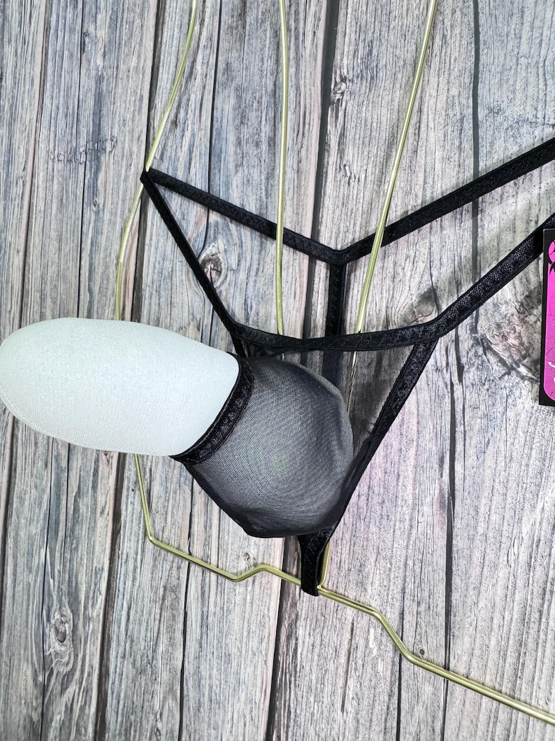 K&ouml;nnte beinhalten: Schwarzes Dessous-Set mit einem transparenten String-Tanga und einem BH-&auml;hnlichen Design. Das Set wird auf einem goldfarbenen Kleiderb&uuml;gel vor einem Holzgrund pr&auml;sentiert. Der Stoff wirkt zart und das Design ist minimalistisch.