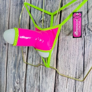 Puede incluir: Un artículo novedoso rosa neón y verde. El artículo es una prenda estilizada en forma de falo con una punta blanca, ribeteada en verde brillante. Está suspendido por un marco de alambre dorado. Una etiqueta con el texto "jovanadesign" es visible.