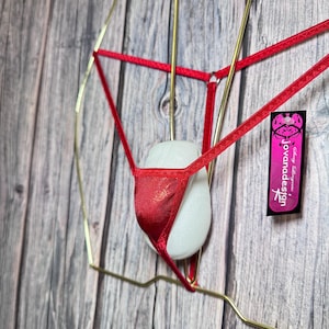 Könnte beinhalten: Ein roter, glänzender String-Tanga wird auf einem goldfarbenen Drahtrahmen vor einem Holzgrund gezeigt. Die Dessous haben einen metallischen Glanz. Ein Etikett mit dem Text "Jovanadesign" ist sichtbar.