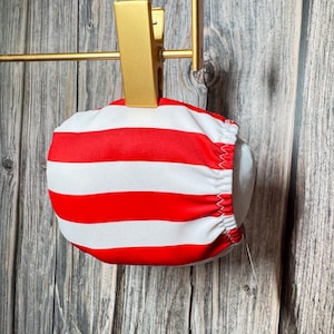 May include: A red and white striped garment, possibly swimwear, is held by a gold-colored metal clip. The fabric is gathered at the edges. The background is a weathered, gray wooden surface.