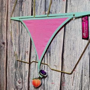 May include: Pink thong underwear with a mint green trim and waistband. A peach-shaped charm and a purple gem are attached to the underwear. The underwear is displayed on a gold-colored wire hanger against a wooden background.