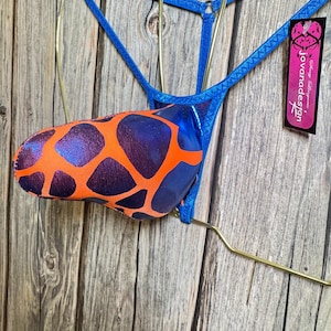 May include: A blue and orange patterned thong with a Joviana Design tag. The thong is hanging on a gold hanger against a wooden background.