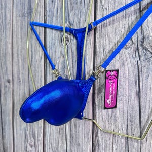 Puede incluir: Un tanga de baño azul metálico con tirantes finos y una parte delantera en forma de bolsa. El traje de baño está colgado de una percha de metal dorado sobre un fondo de madera. Una etiqueta con el texto "jovanadesign" está adjunta.