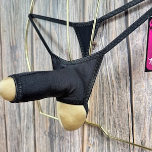 May include: Black thong underwear with a gold trim. The underwear is hanging on a gold hanger against a wooden background.