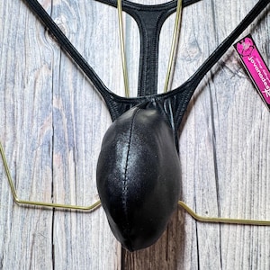 May include: Black thong underwear with a pouch, hanging on a gold-colored hanger. The underwear has a smooth, shiny texture. A tag with pink and white text is visible on the right side.