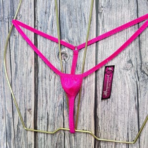 May include: A bright pink thong, displayed on a gold-colored hanger against a wooden background. The thong features a delicate design with thin straps and a small pouch. A tag with the text "jovanadesign" is attached.