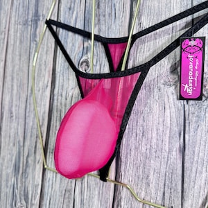 May include: A hot pink mesh thong with black trim, displayed on a gold-colored hanger. The lingerie features a pouch and a tag with the text "jovanadesign". The background is a wooden surface.