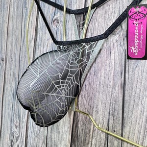 Könnte beinhalten: Schwarze und silberne Dessous mit Spinnennetz-Muster und schwarzem Besatz. Die Dessous werden an einem goldfarbenen Kleiderbügel präsentiert. Ein pinkfarbenes Etikett mit dem Text "jovanadesign" ist angebracht.