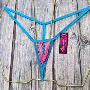 Könnte beinhalten: Ein türkisfarbener String mit einem rosafarbenen Spitzeneinsatz. Die Dessous werden auf einem goldfarbenen Kleiderbügel vor einem Holzmaserungshintergrund präsentiert. Ein kleines Etikett mit dem Text "Jovanda Design" ist angebracht.