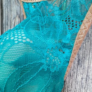 May include: Close-up of a teal lace garment with a floral pattern. The edges are trimmed with a thin, gold-colored material. The background is a weathered, gray wooden surface.