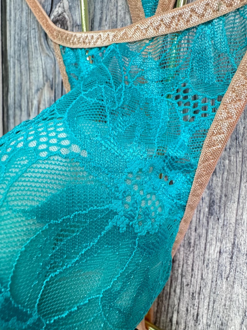 May include: Close-up of a teal lace garment with a floral pattern. The edges are trimmed with a thin, gold-colored material. The background is a weathered, gray wooden surface.