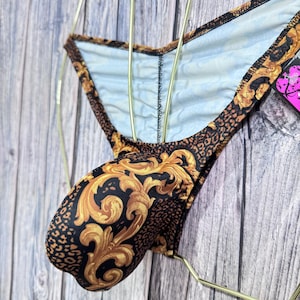 May include: A pair of black and gold patterned underwear. The fabric features a baroque design in gold against a black background, with leopard print accents. The underwear is displayed on a gold hanger.