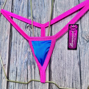 May include: A vibrant pink and blue thong bikini bottom. The bikini bottom features a bright pink trim and straps, with a blue triangle front. A tag with the text "jovanadesign" is attached.