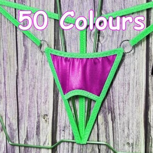 Puede incluir: Un tanga verde y morado con el texto "50 Colores" en blanco. El tanga tiene forma de triángulo con un centro morado y ribete verde. El fondo es una superficie de madera.